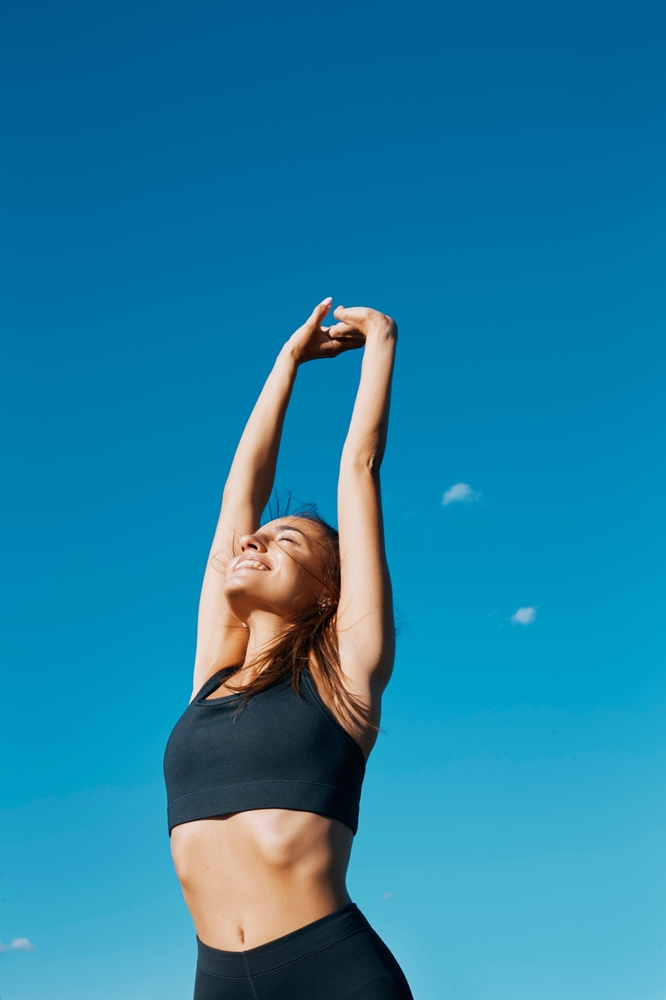 A smiling athletic woman stretching under a blue sky, showcasing the joint recovery benefits of BPC-157 peptide therapy at Relive Brentwood.
